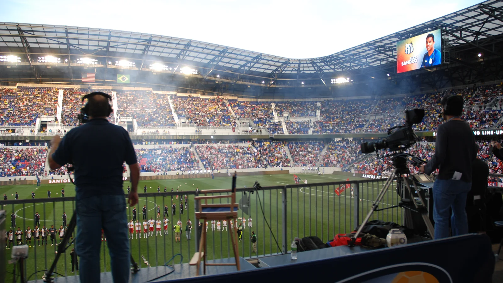 Cámara de televisión y operador frente al terreno de juego de un estadio de fútbol lleno.